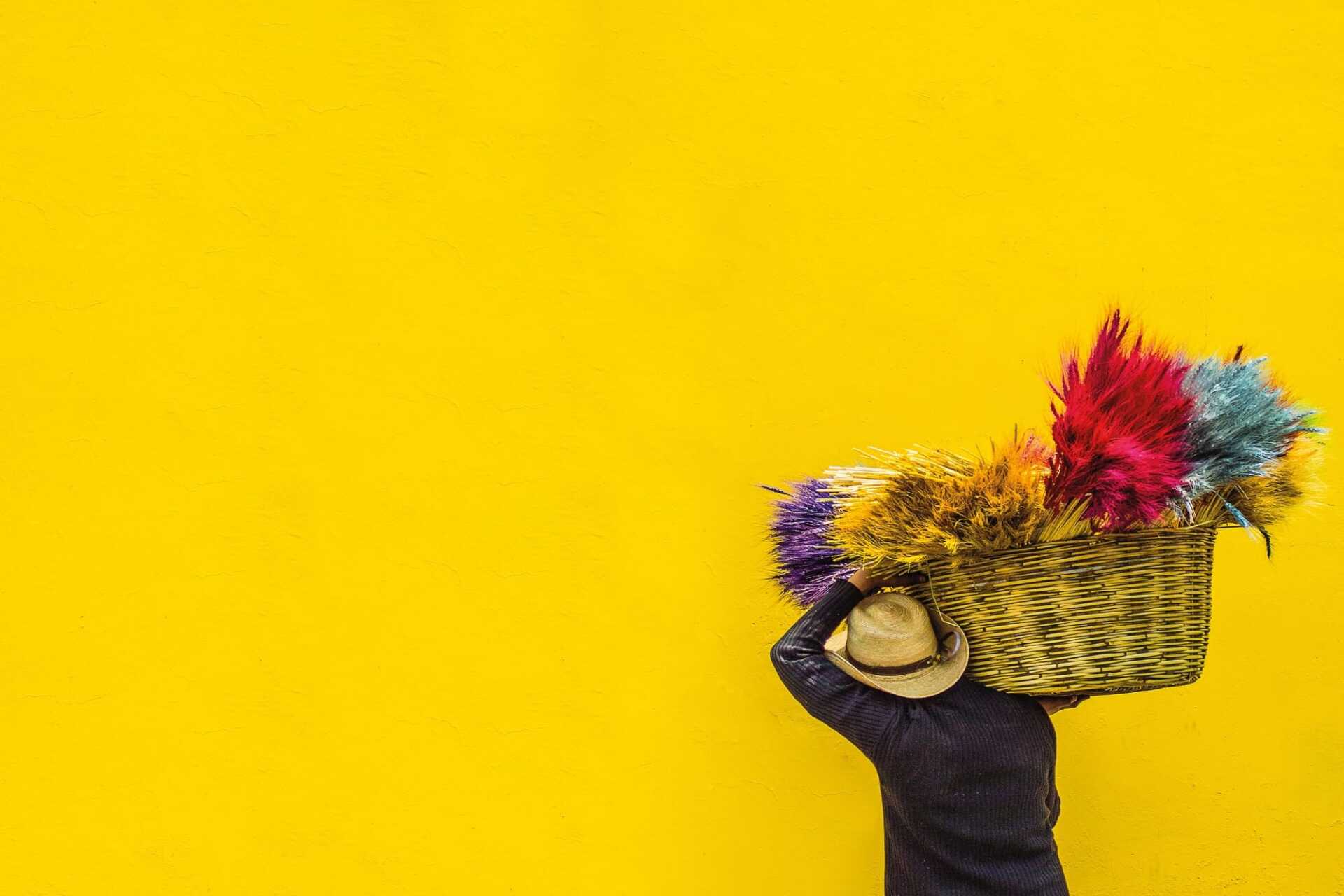 Man facing away from us, with hat against a yellow wall. He's carrying a basket of brightly coloured flowers.