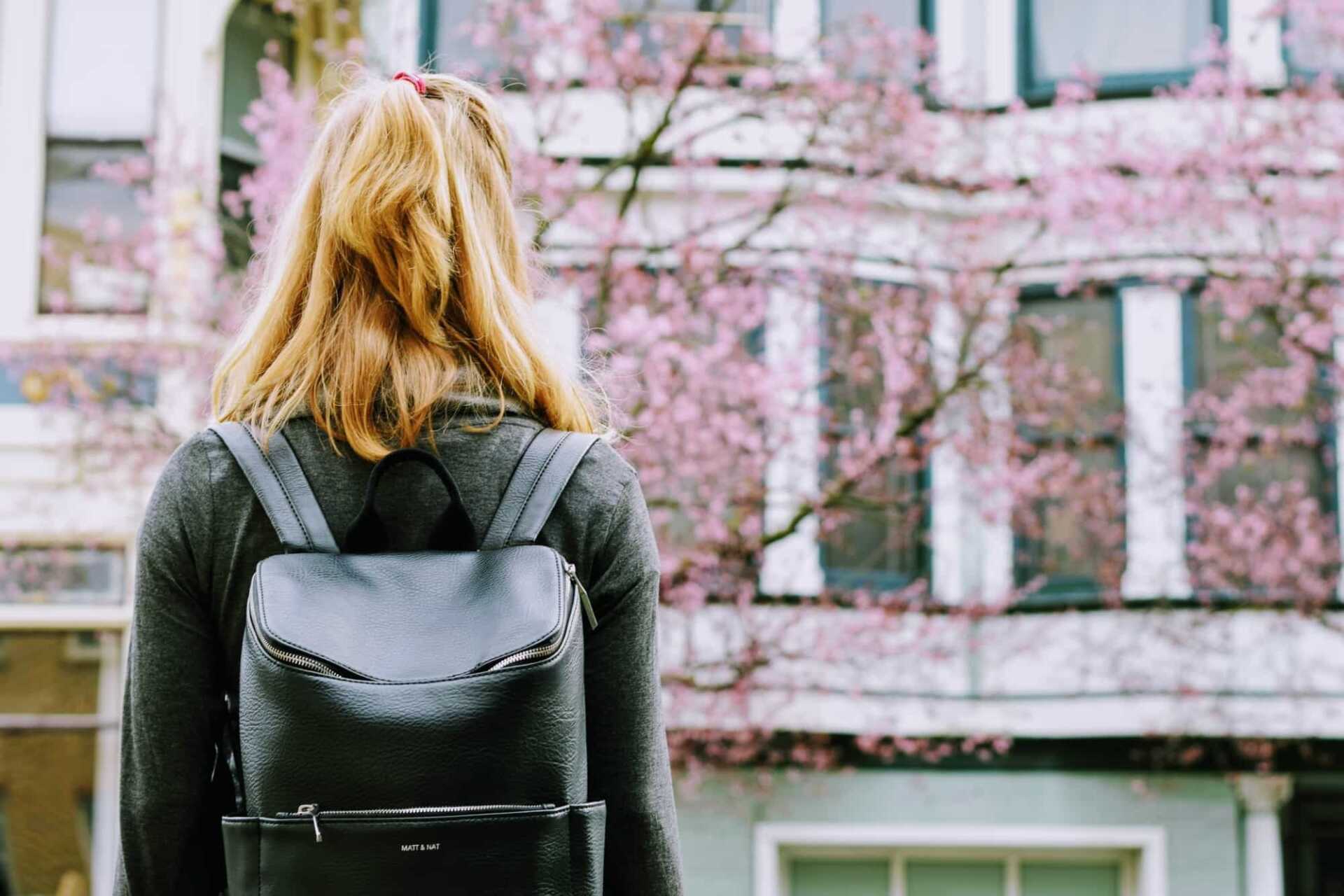 Blonde woman wearing a rucksack, seen from behind standing in front of a building and a cherry blossom tree