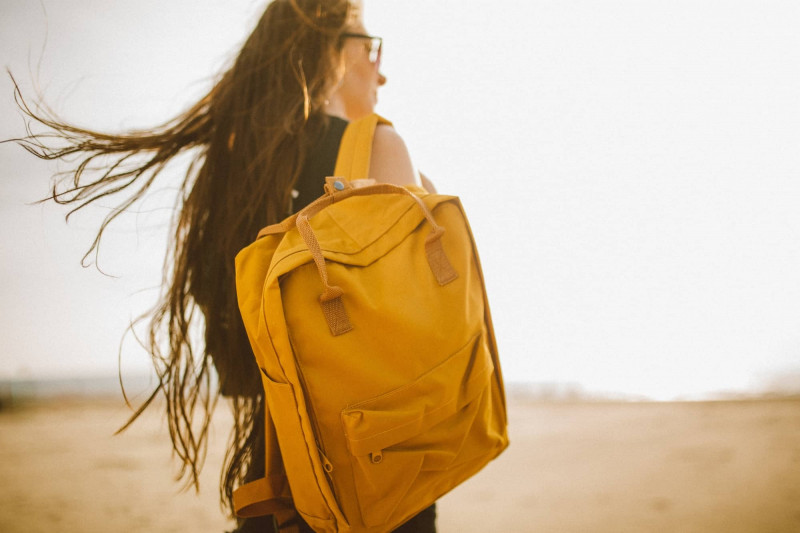 Woman with dark hair standing on a beach. Her hair is blowing in the wind, and she has a yellow bag over her right shoulder.