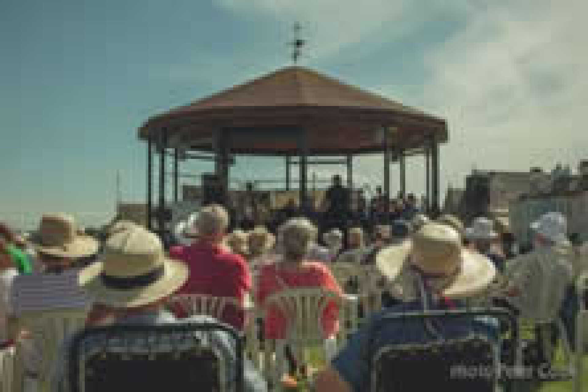 Sun-hat wearing audience watching a band perform on a bandstand