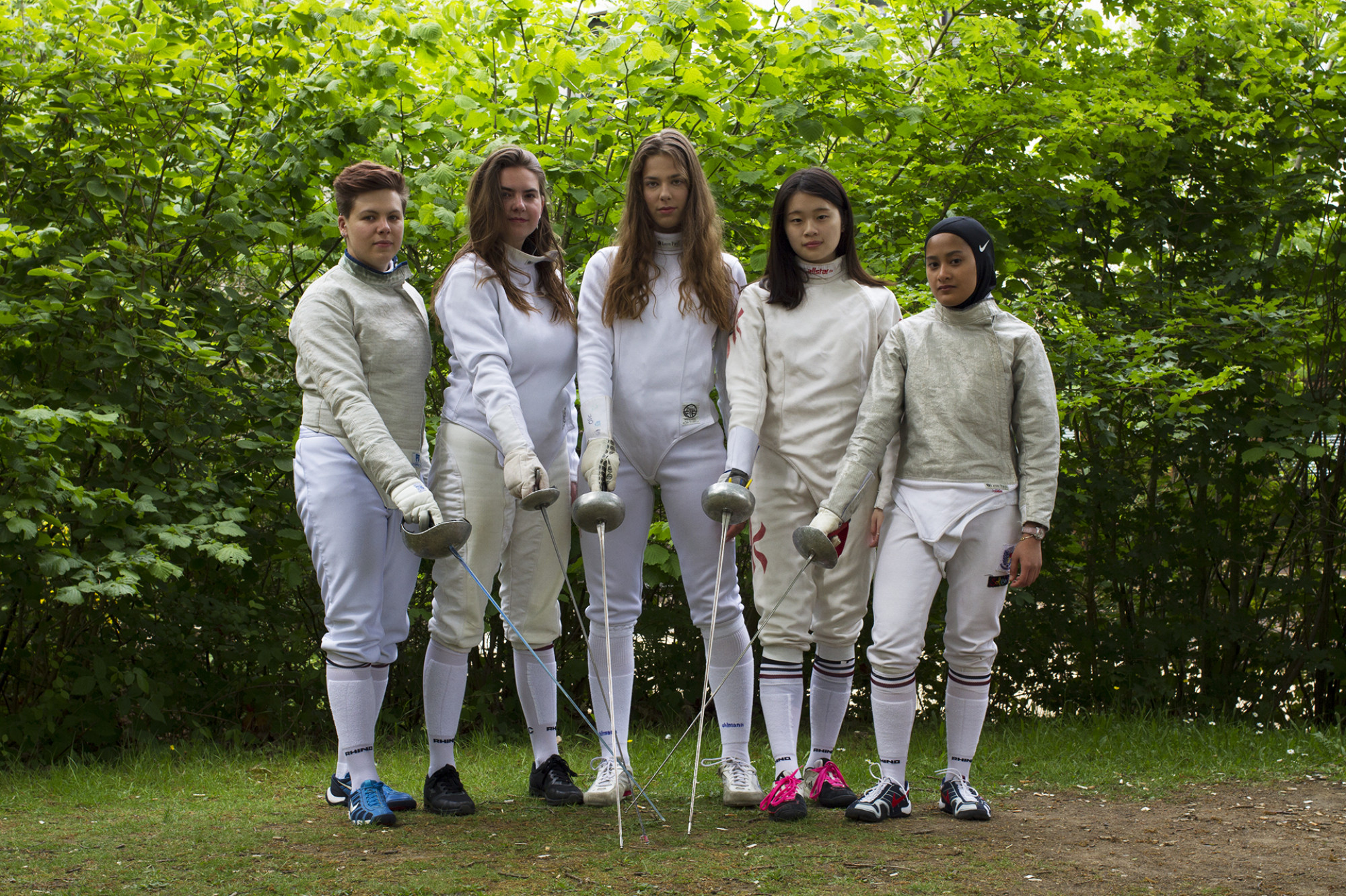 Women's fencing team stood together in wooded area in full sports kit holding swords toward ground in a line
