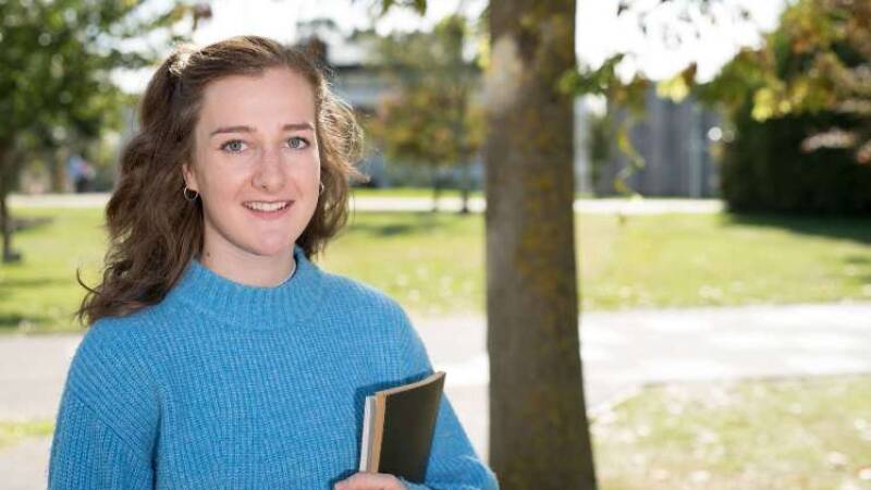 Becky wears a blue sweater and holds a black notebook. She has brown hair and is smiling to camera.