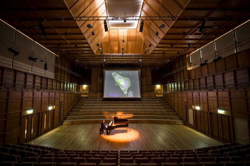 Pianist in Colyer-Fergusson Concert hall plays in front of screen showing cells