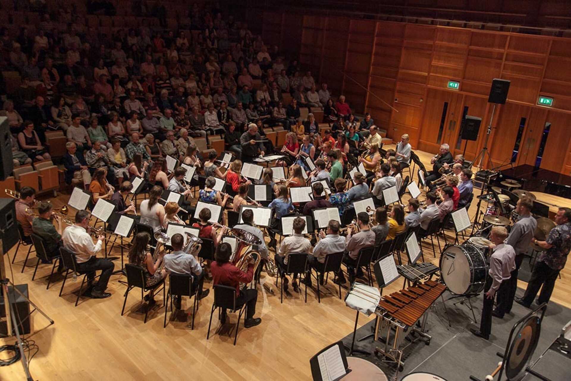 View from behind massed ranks of concert band musicians playing in front of an audience in a concert-hall