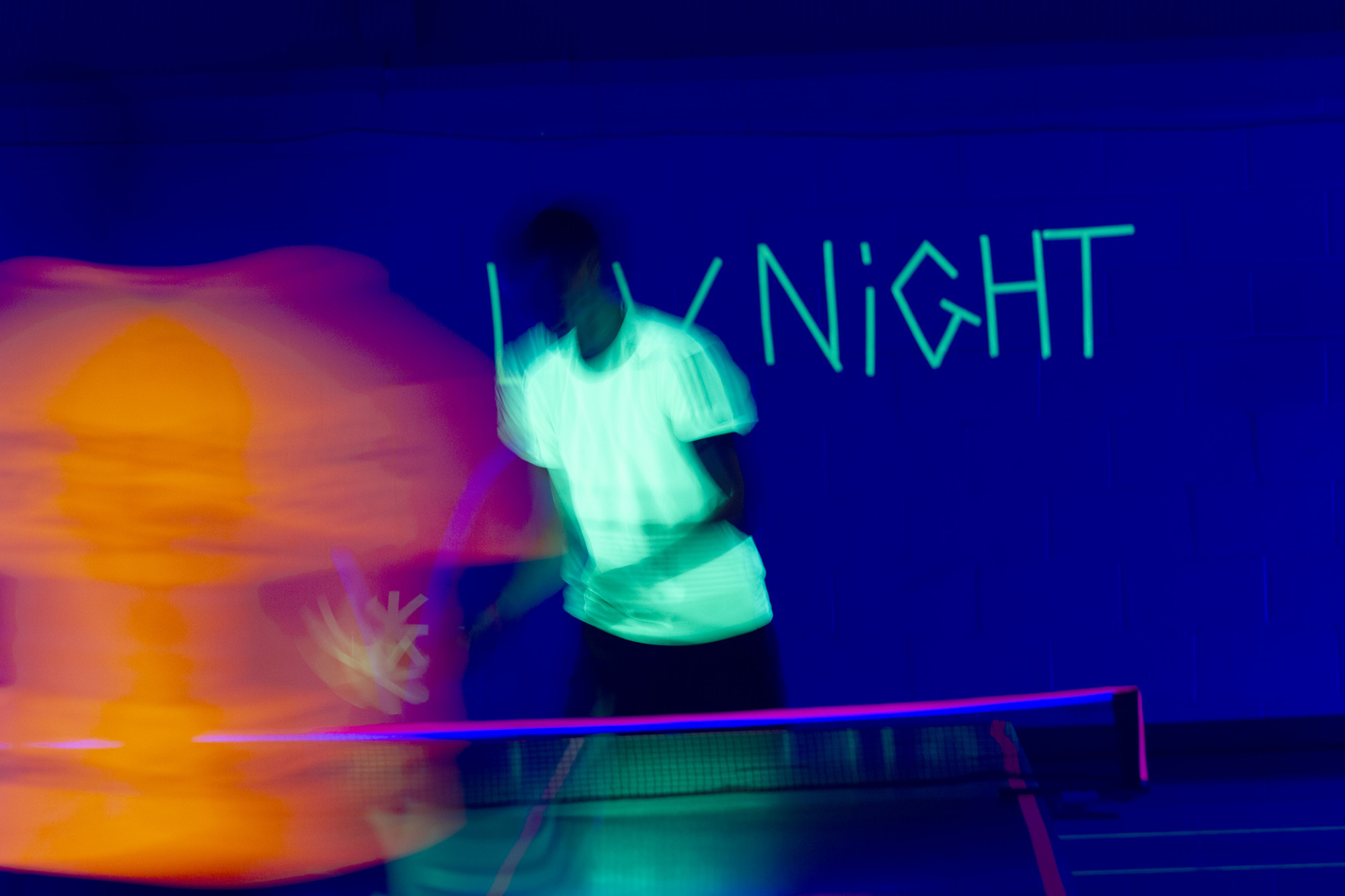 Blurry action photo of two people playing table tennis in the dark with UV lights