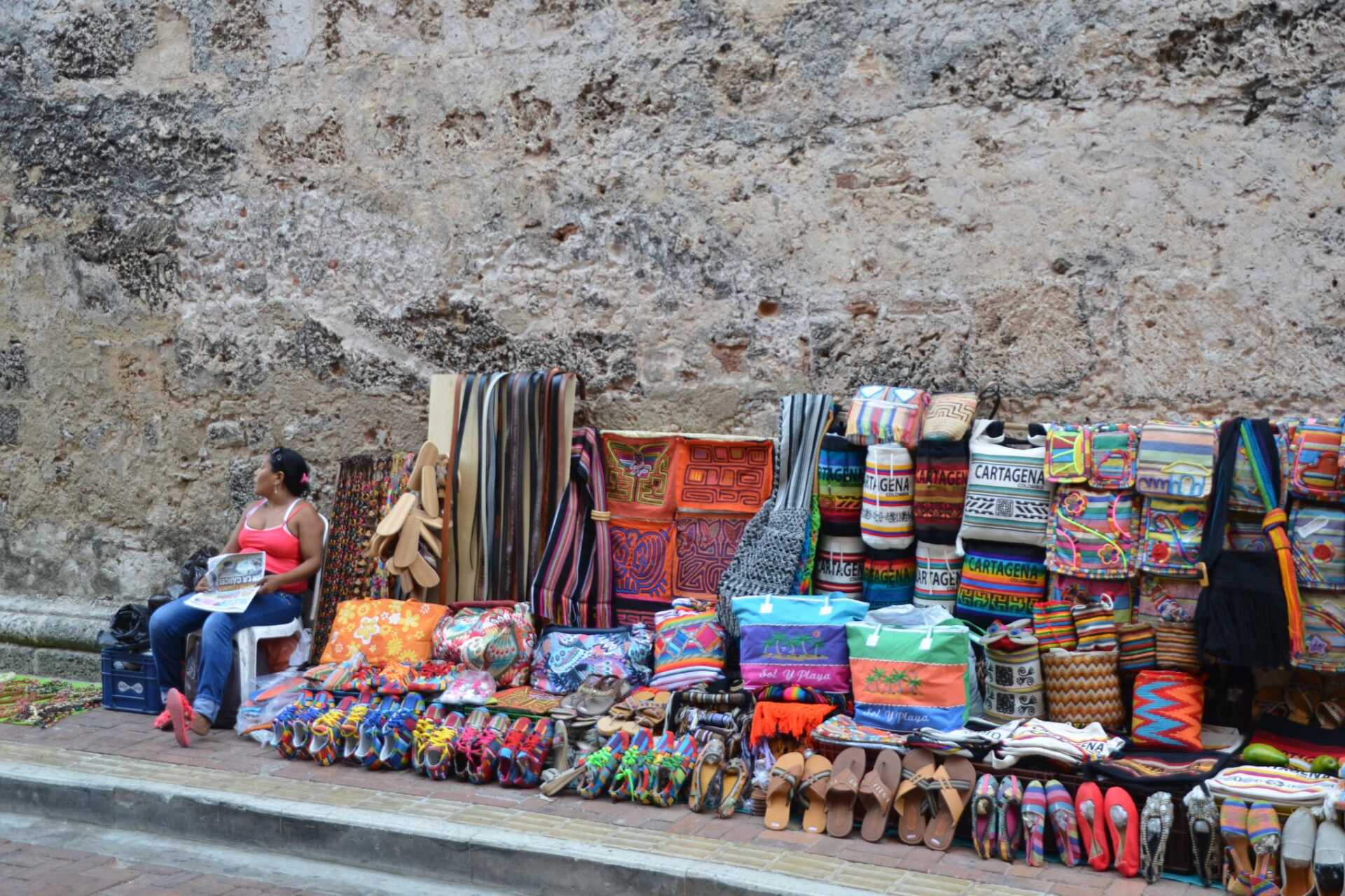 A woman sitting on a chair on a street with rows of colourful crafts lined up against a wall on sale