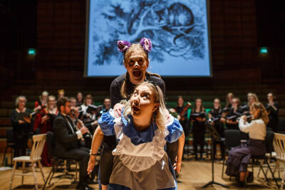 One female singer giving another a piggy-pack wearing Alice in Wonderland costumes, a group of musicans playing in the back