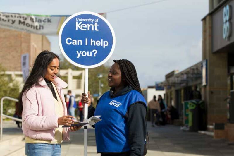 Two female students, one with a University of Kent sign with 'Can I help you?' on it