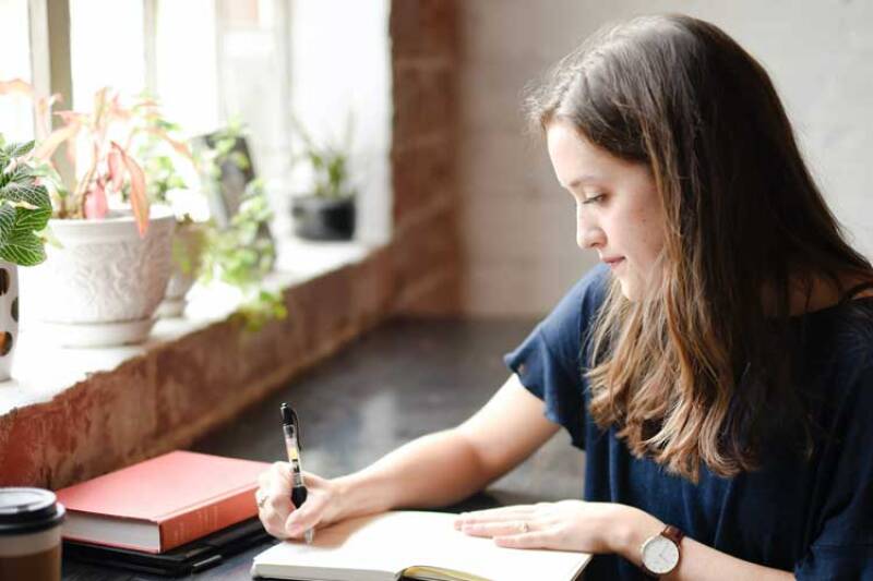 Young woman, long brown hair, writing at a desk Image by Hannah Olinger Unsplash