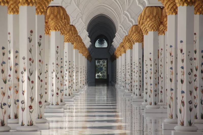 A hallway in a mosque in Abu Dhabi, with ornate pillars either side. The floor, columns and ceiling are white.