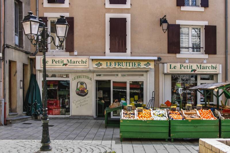 A town square with displays of vegetables outside a shop with Le Fruitier written above the door.
