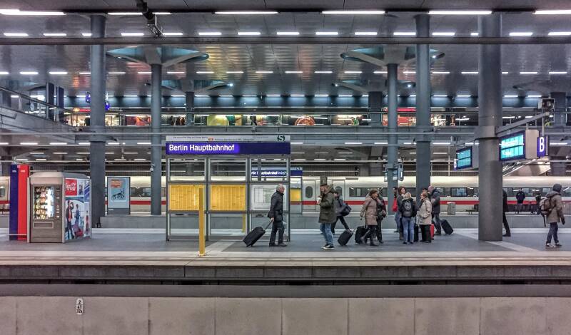 Image of a station platform, with people and suitcases. The blue and white station sign says Berlin Hauptbanhof.