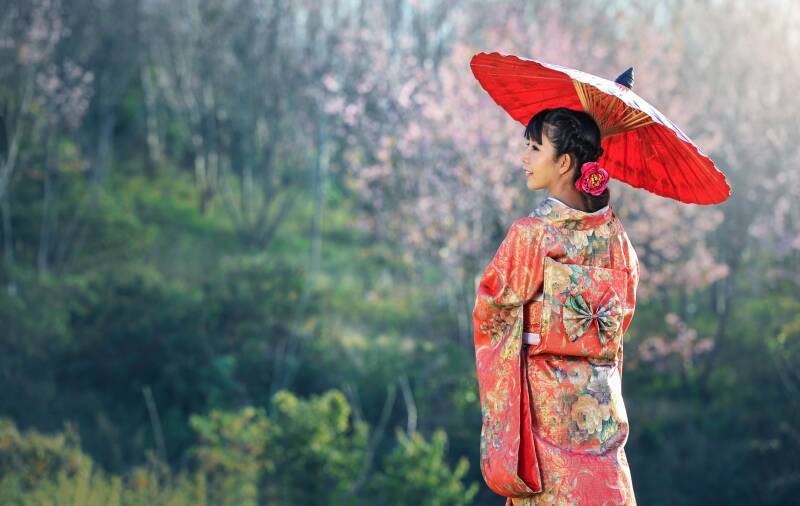 A girl in a red floral kimono faces away from the camera, looking over her left shoulder. She carries a red parasol.