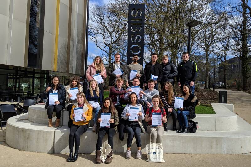 Students sat outside the Sibson Building holding up certificates.