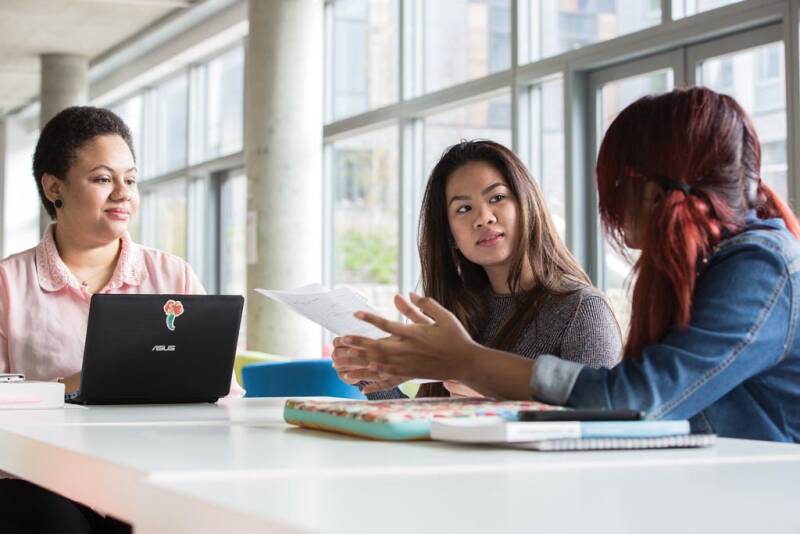 Three women sitting at a table talking with a laptop and notebooks.