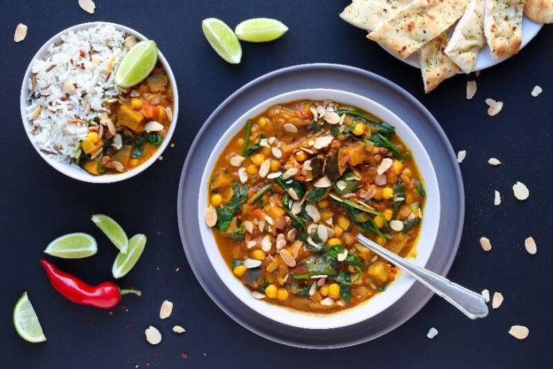 A bowl of vegan curry on a blue background with side dishes of coleslaw and naan bread to the left and right.