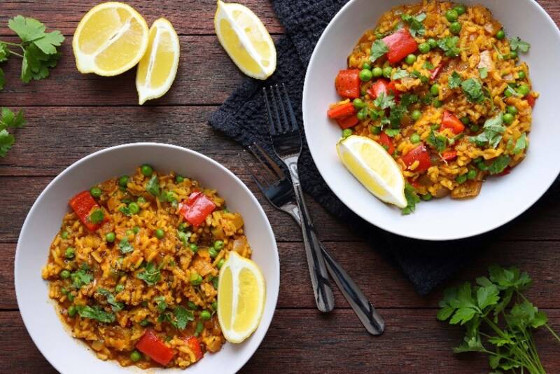 Two bowls of spanish paella are diangonally positioned on a wooden tbale with two forks.