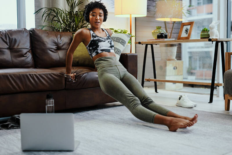 A lady is in her house, working out on her sofa whilst listening or watching a class.