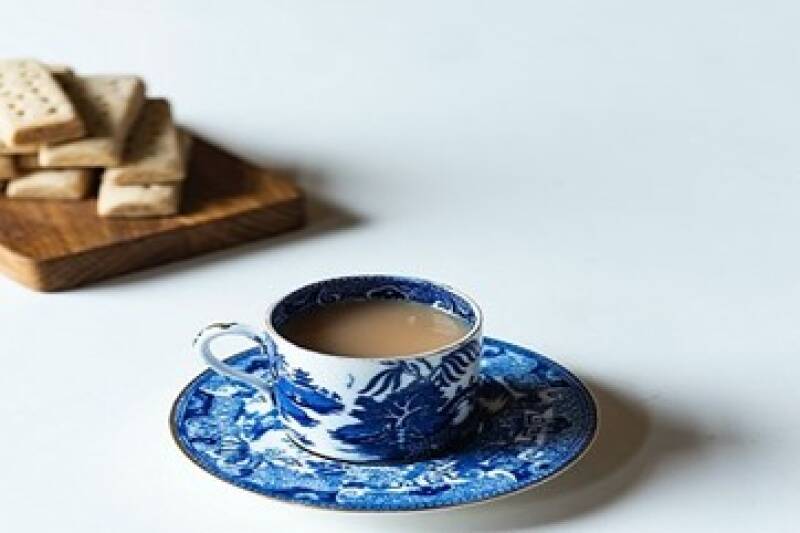 A blue and white flowery tea cup with a wooden board of biscuits blurred and partially shown on a white background