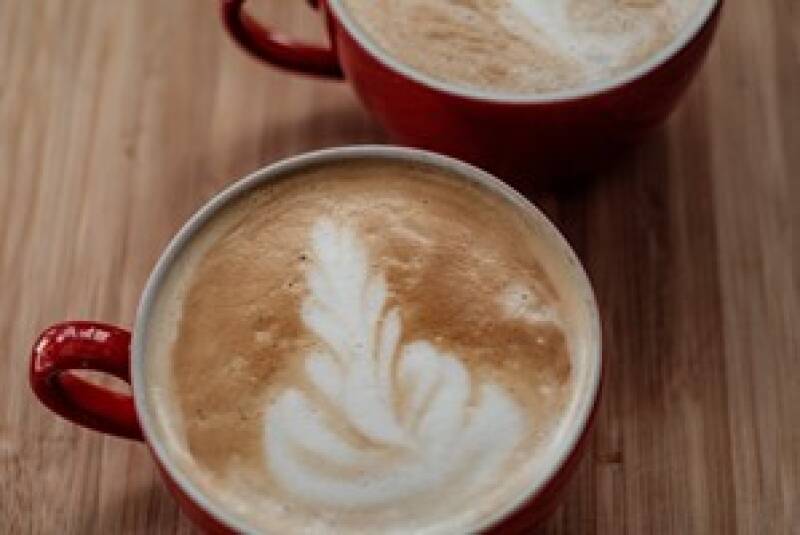 Two red cups zoomed in on to show the leaf design in the coffee. Placed on a wooden table.
