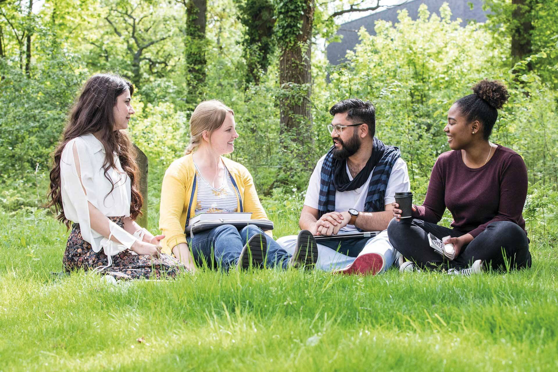 Male and three female students of mixed ethnicity chatting in a green space on campus on a sunny day