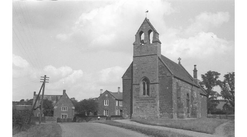 Black and white photograph of a stone church on a quiet village road. In the background are two brick houses.