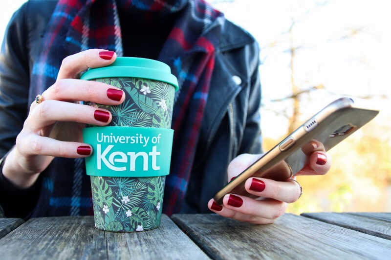 A person holding a Kent branded reusable coffee cup in their right hand whilst using their phone in their right hand