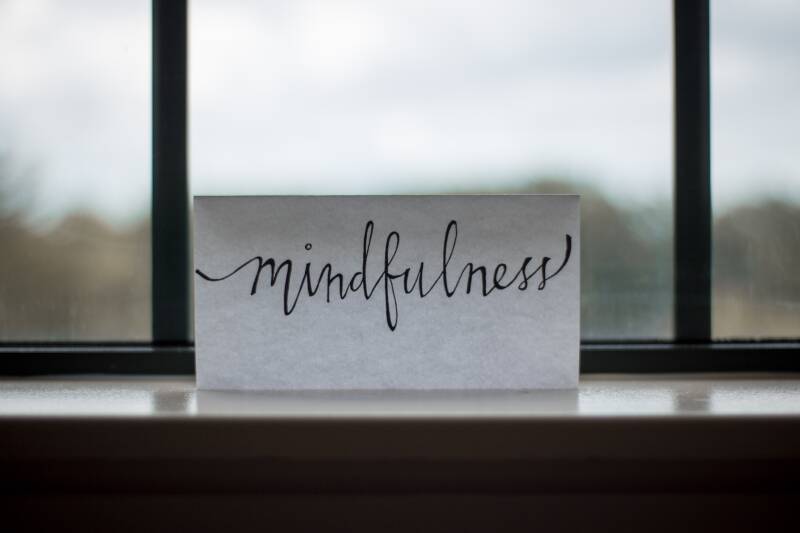 Piece of paper with 'Mindfulness' written in italics, standing on a surface with a big window in the background