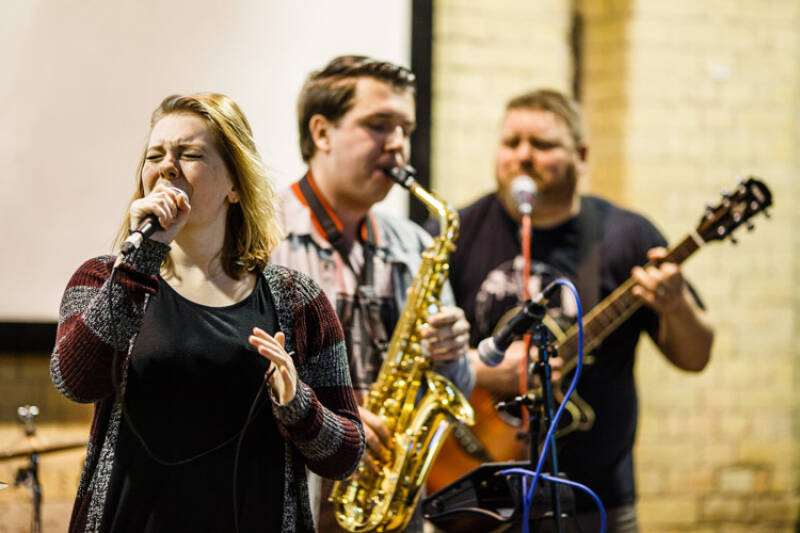 A female student is singing into a microphone, with two male students in the background playing the saxophone and guitar