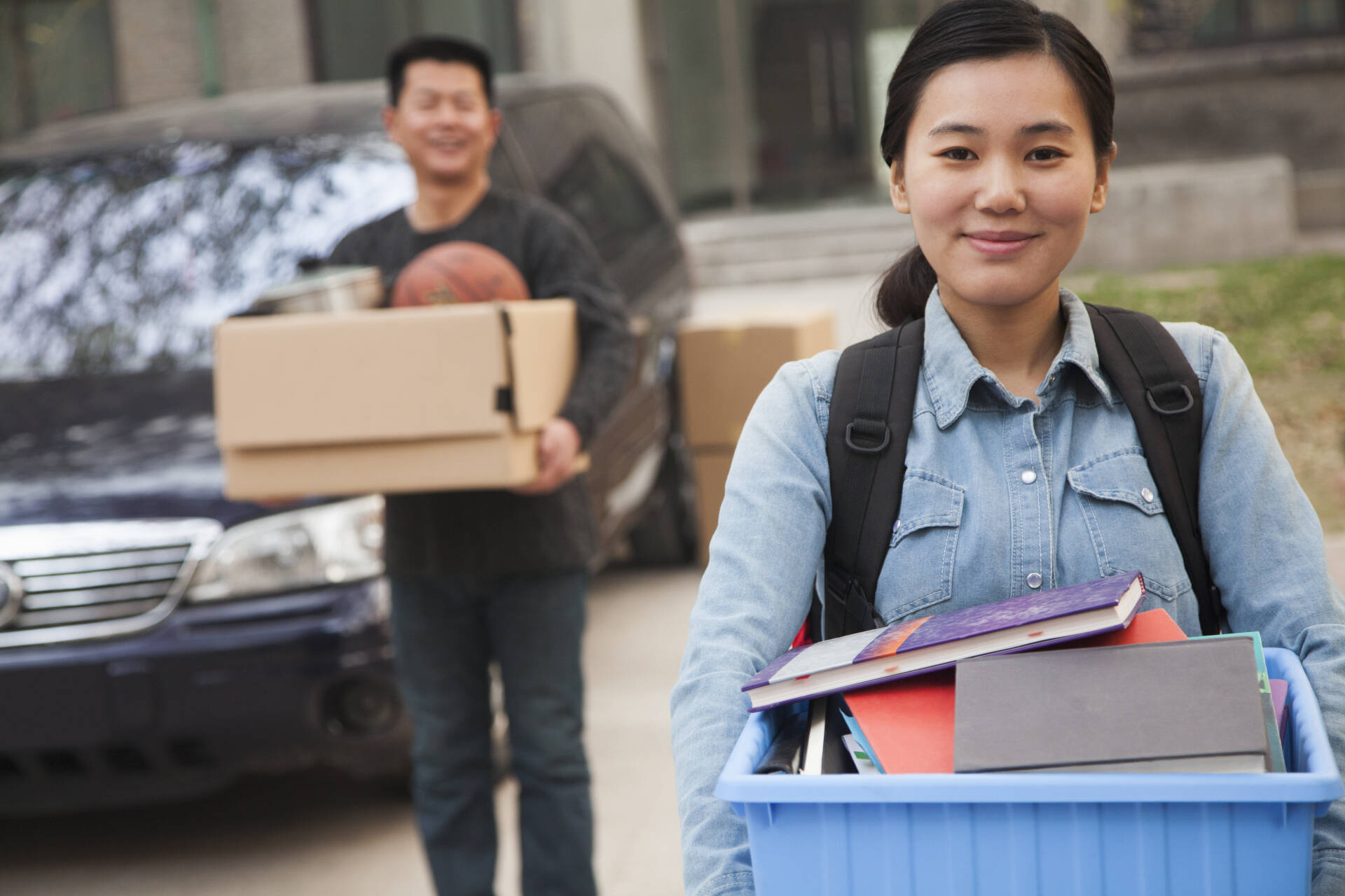 Female student carrying a box of books with father carrying another box from a car in the background