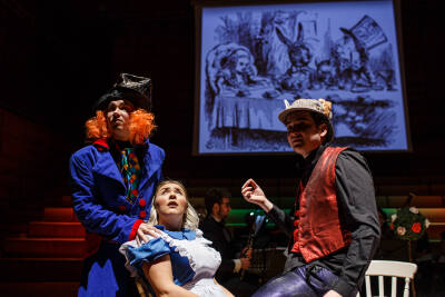 Three costumed performers on stage; a proection screen in the background with an illustration from 'Alice in Wonderland'