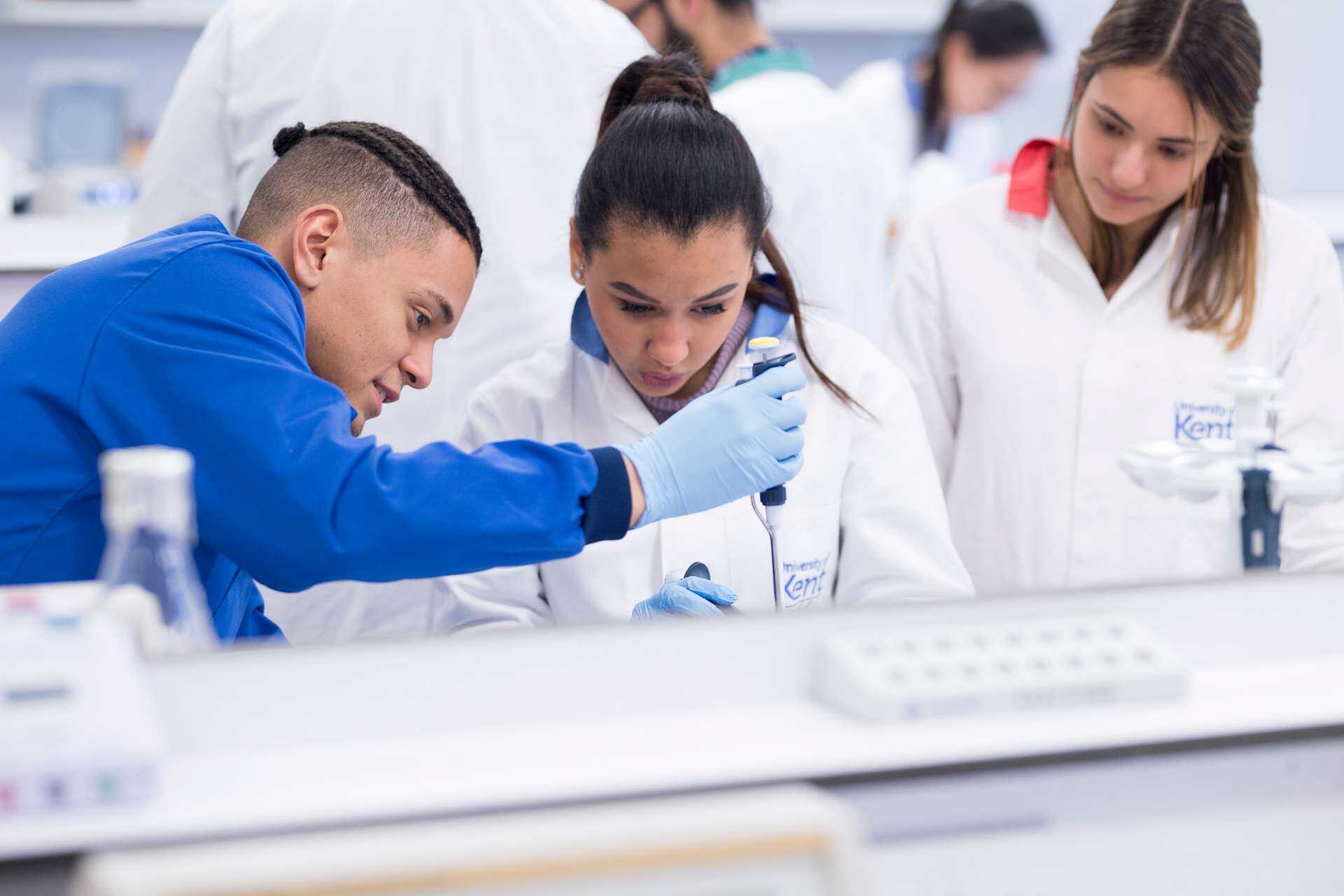 Two students using a pipette in an experiment whilst another female student looks on in the background