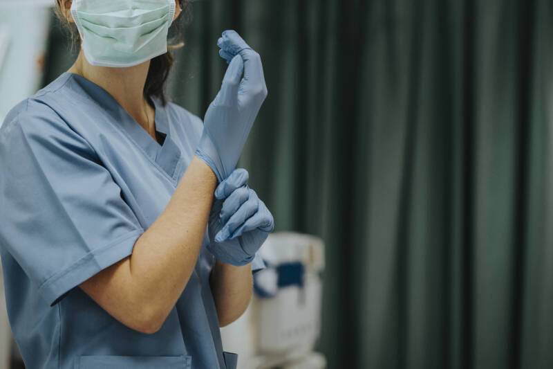 Woman dressed in blue scrubs and wearing a mask pulls on a pair of surgical gloves