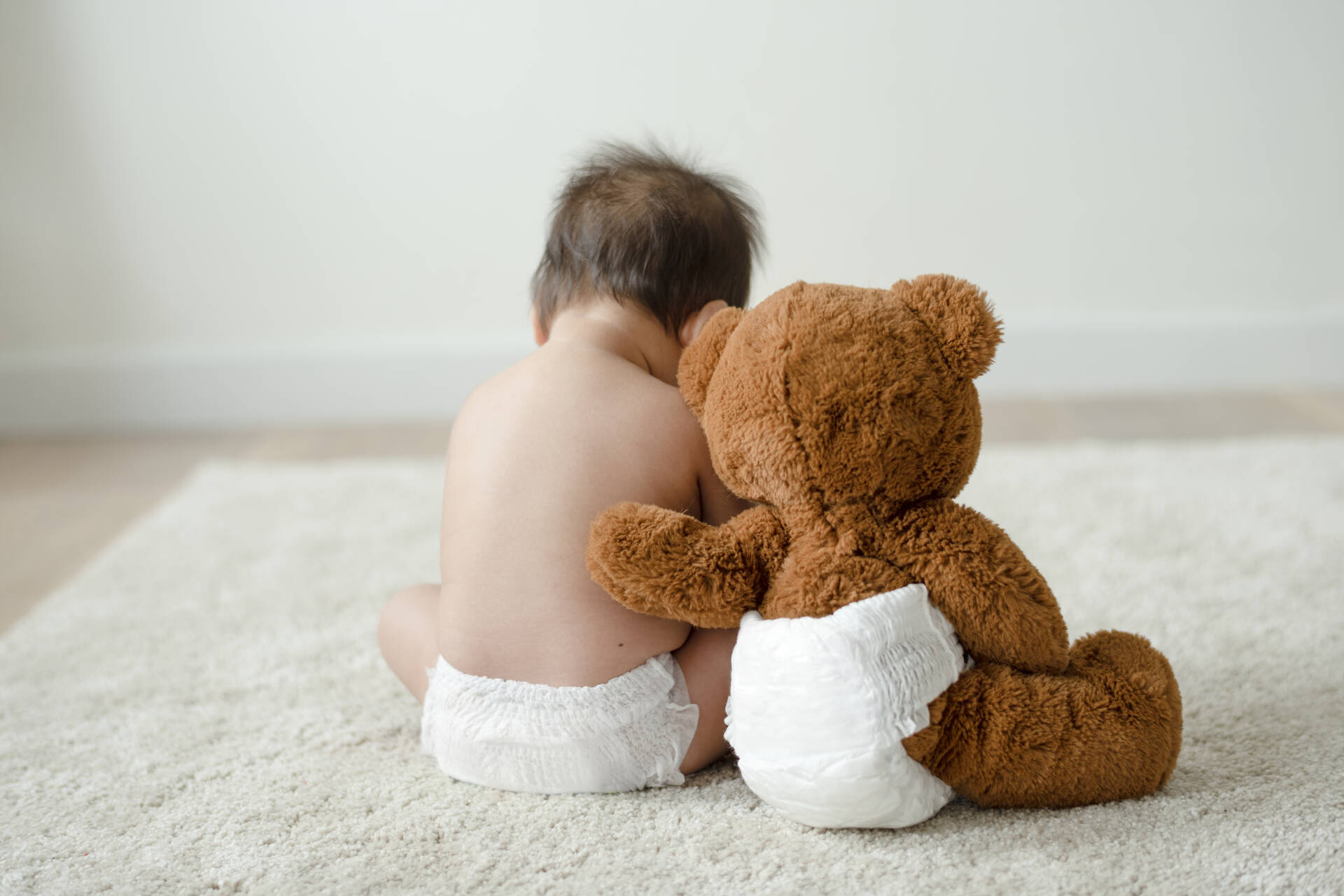 A baby sits beside a teddy bear on a carpet. The teddy bear's arm is on the child's back
