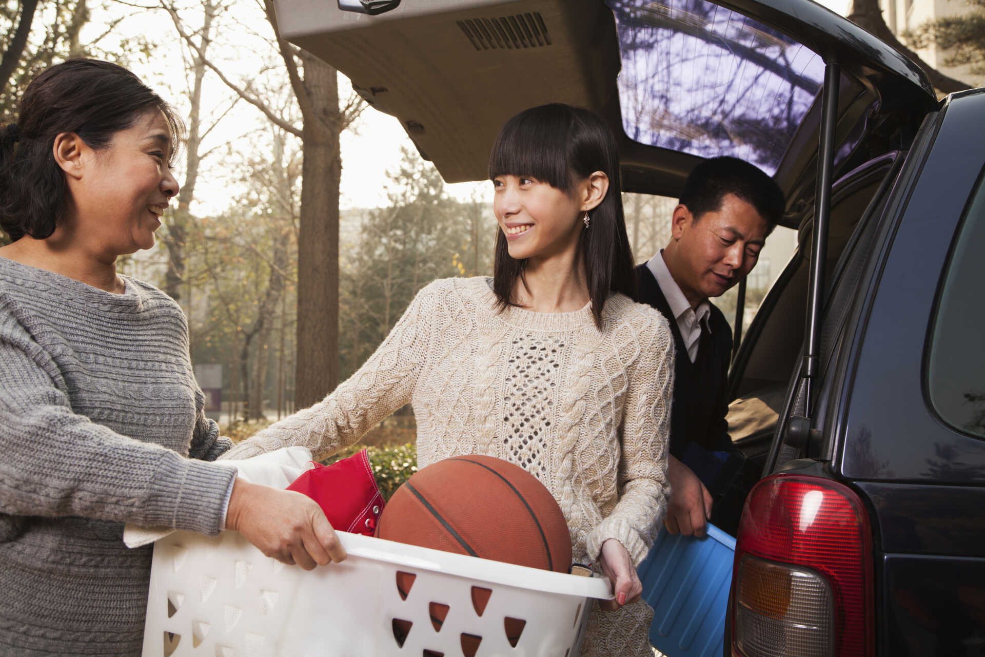 Female student with family carrying boxes of books with father carrying another box from a car in the background