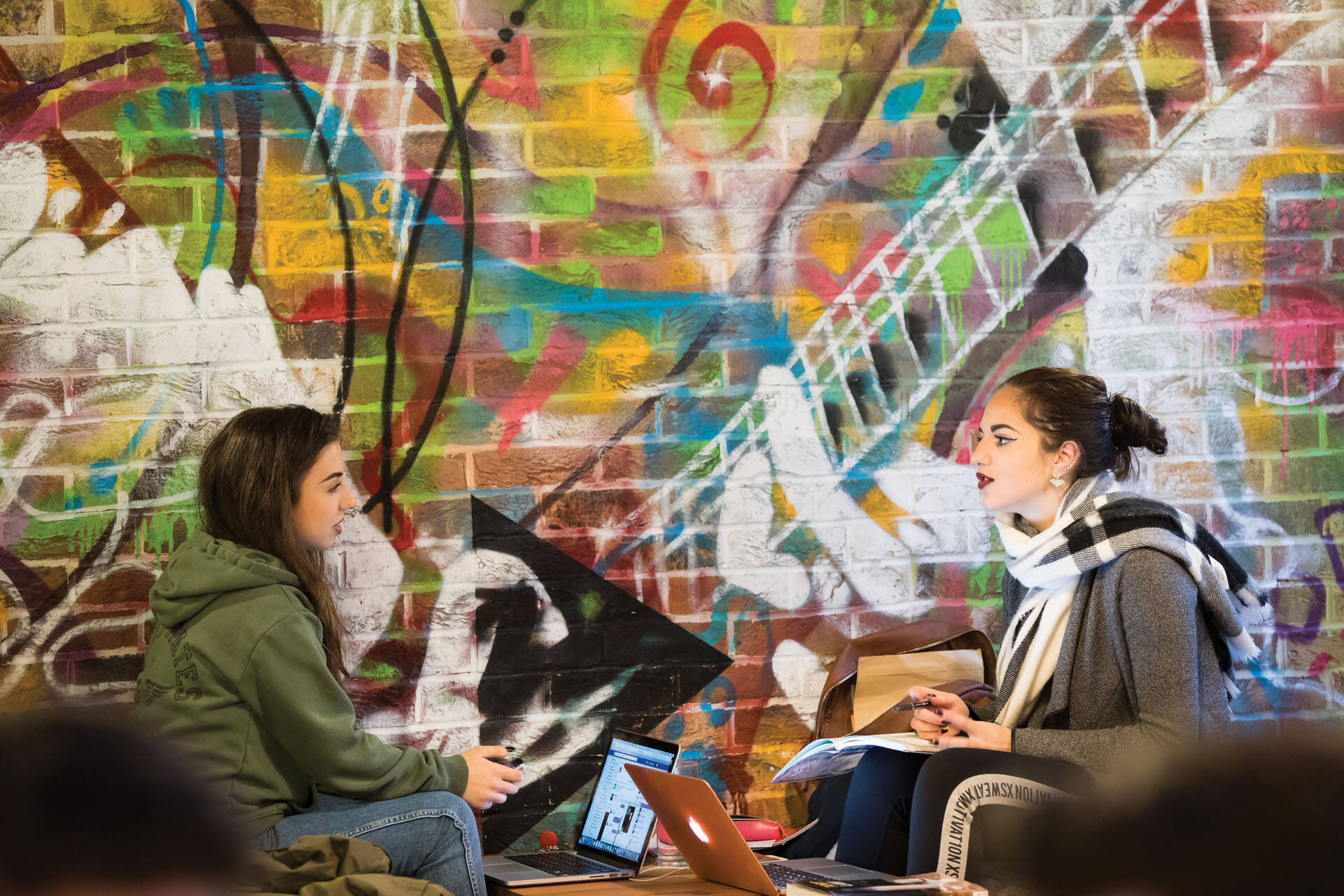 Two female students talking with a graffetti wall in the background.
