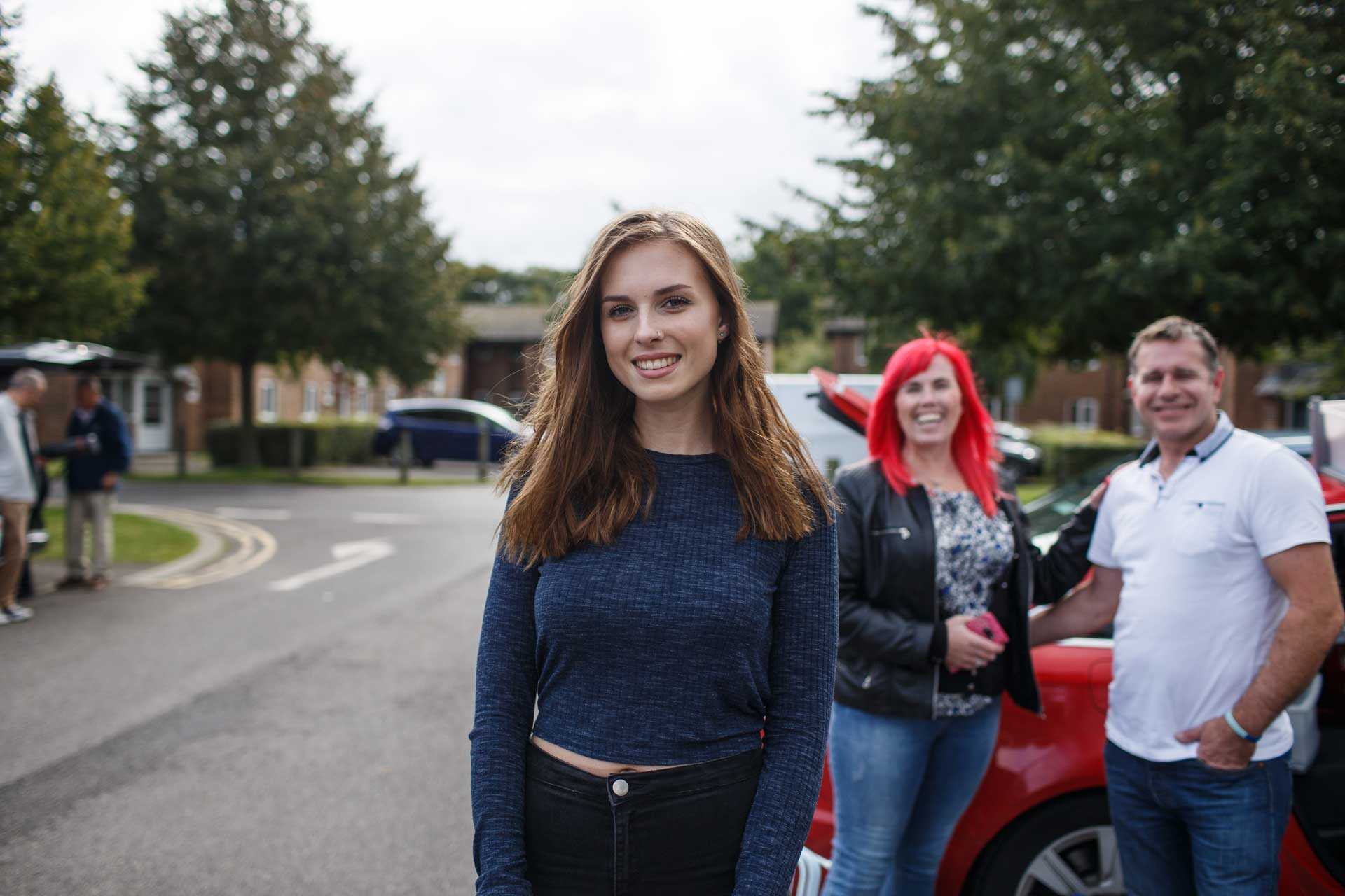 Female student smiling at camera with parents and car in the background also happy