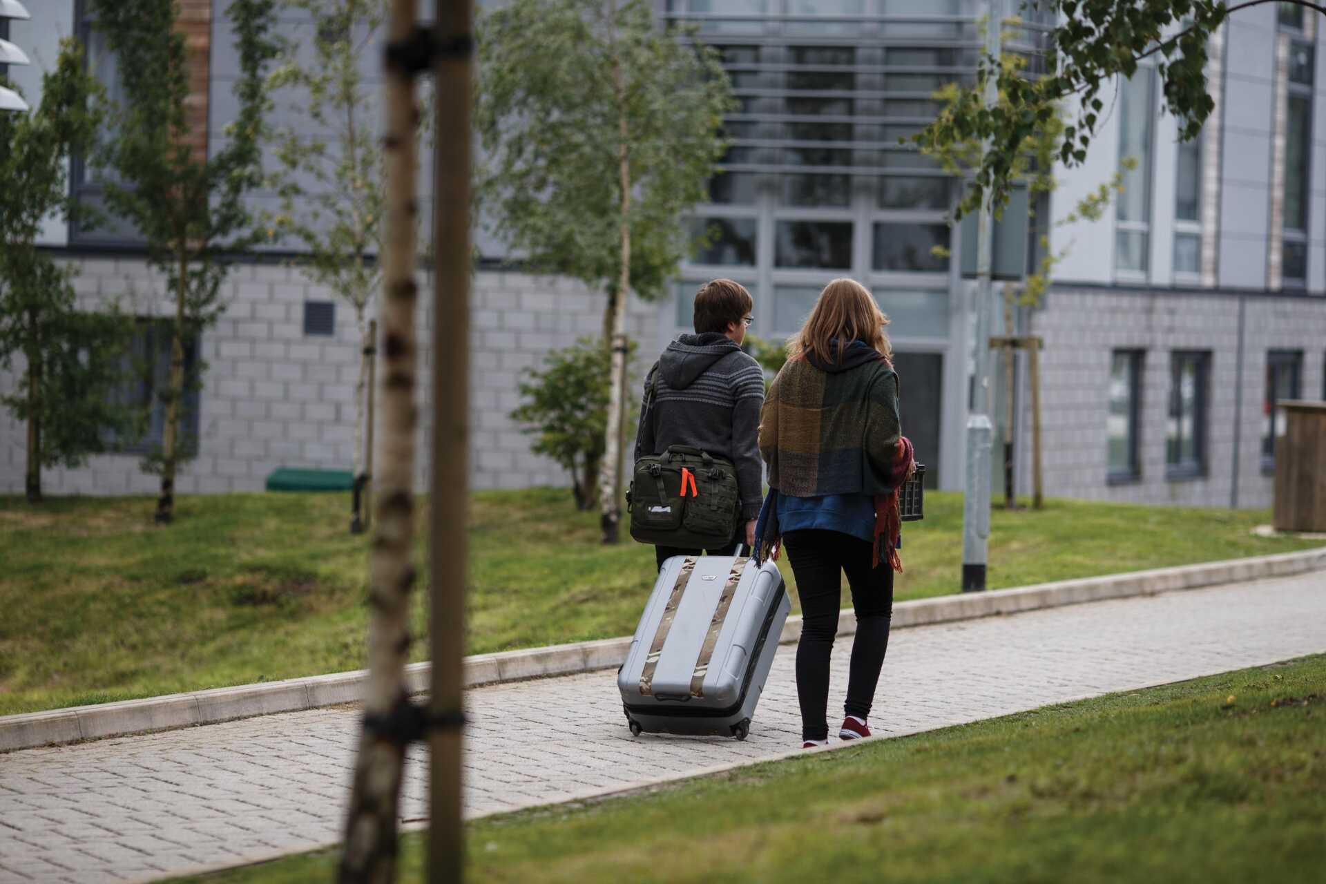 Two people walking out of a building, pulling a suitcase and carrying bags