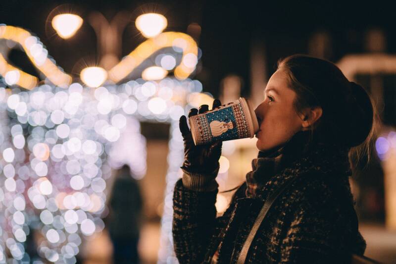 Female wearing scarf and gloves drinking a coffee outside with Christmas lights in the background