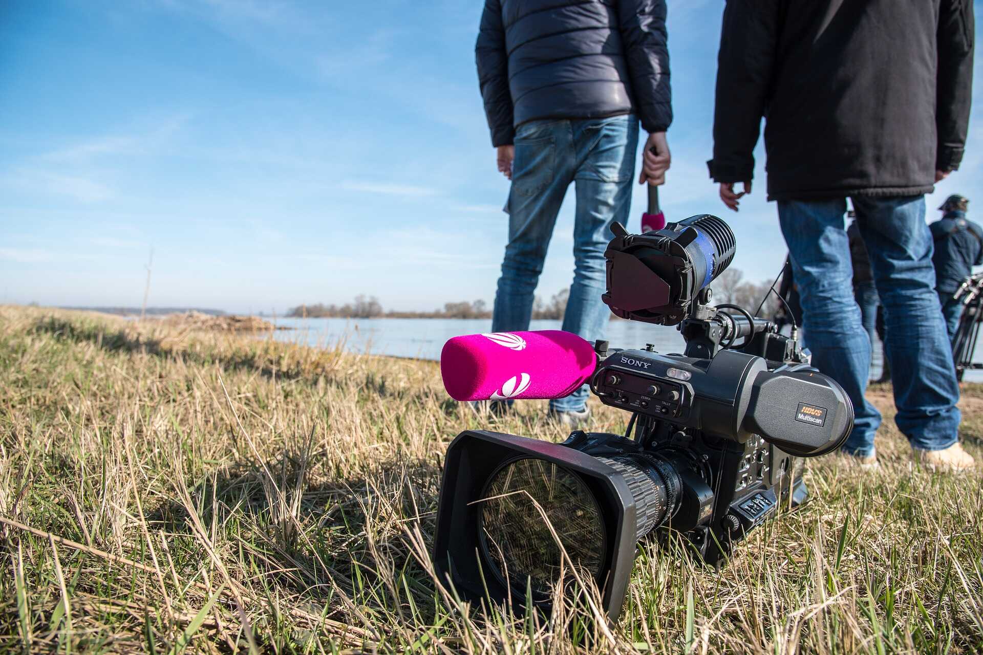 A camera in the foreground, lays on the grass. In the background, a group of people including one holding a microphone