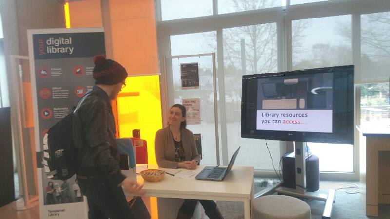 A woman sitting behind a table with a stand-up banner and digital screen. A man has approached the table