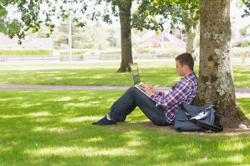 Male student typing on laptop sitting outside on grass