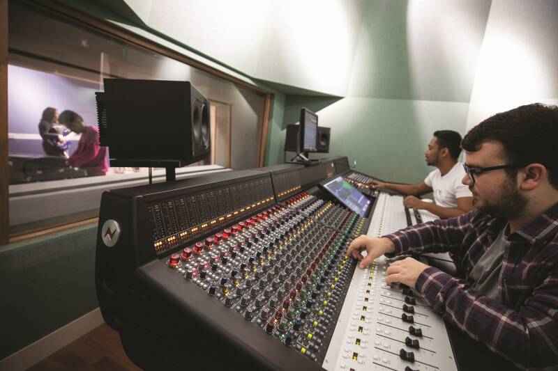 Two students sit at the mixing console. Another student is visible in the studio behind a window.