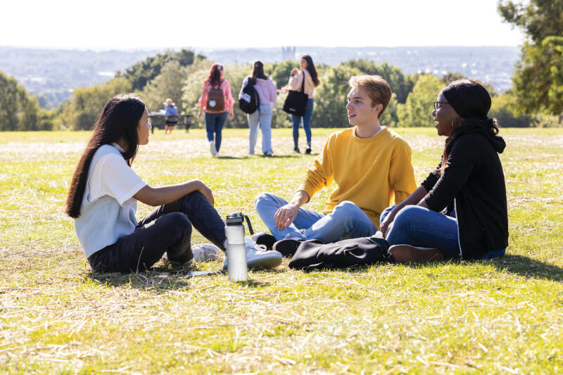 A group of three students sit chatting on the grass whilst a further group of three students walks away across the grass