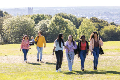 A group of four smiling students, followed by a further two students, walking across a field on campus on a sunny summer day