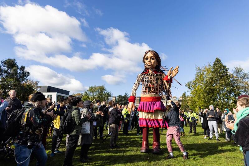 Little Amal puppet on Canterbury campus, surrounded by crowns of people looking. Puppet several times life size.