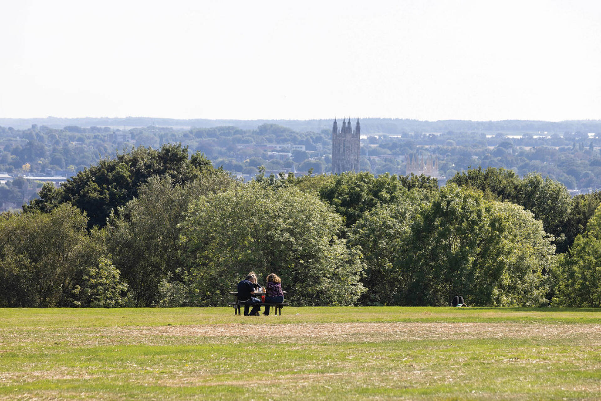 People seated on a bench in the background, with greenery around and Canterbury Cathedral and trees in the background.