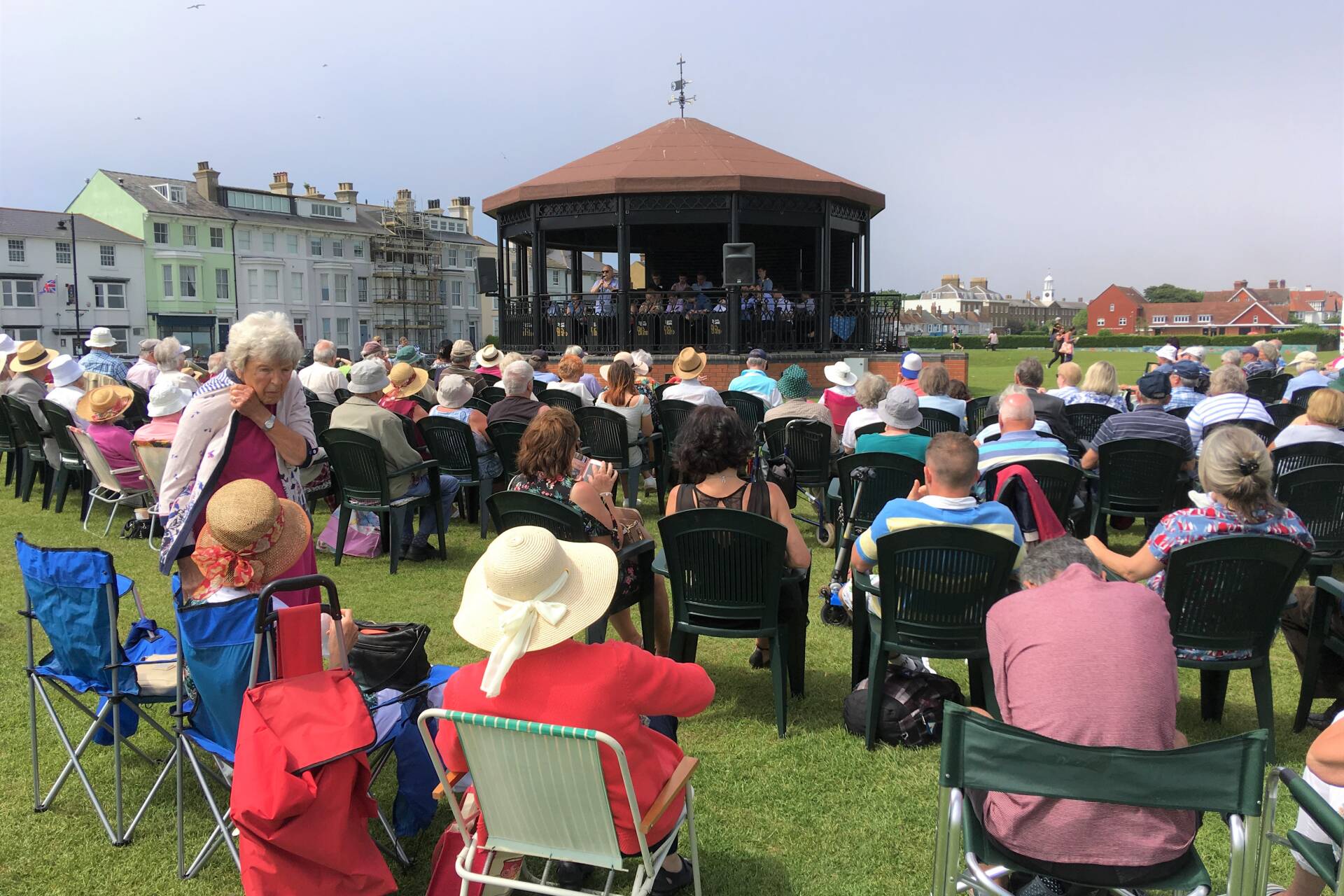 Musicians playing at a bandstand on a greensward, audience looking on