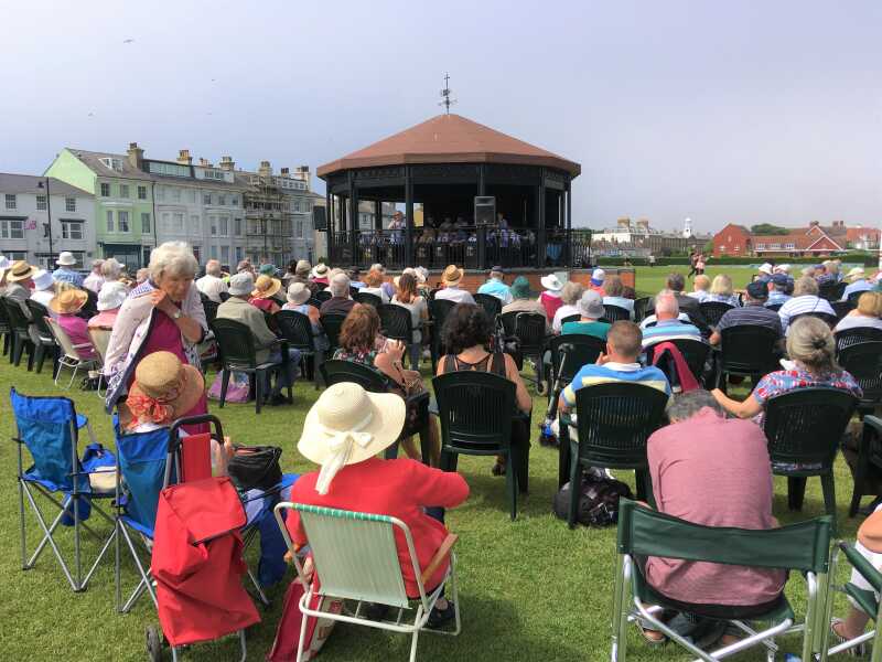 Audience in deck-chairs on a green listening to a band on a bandstand