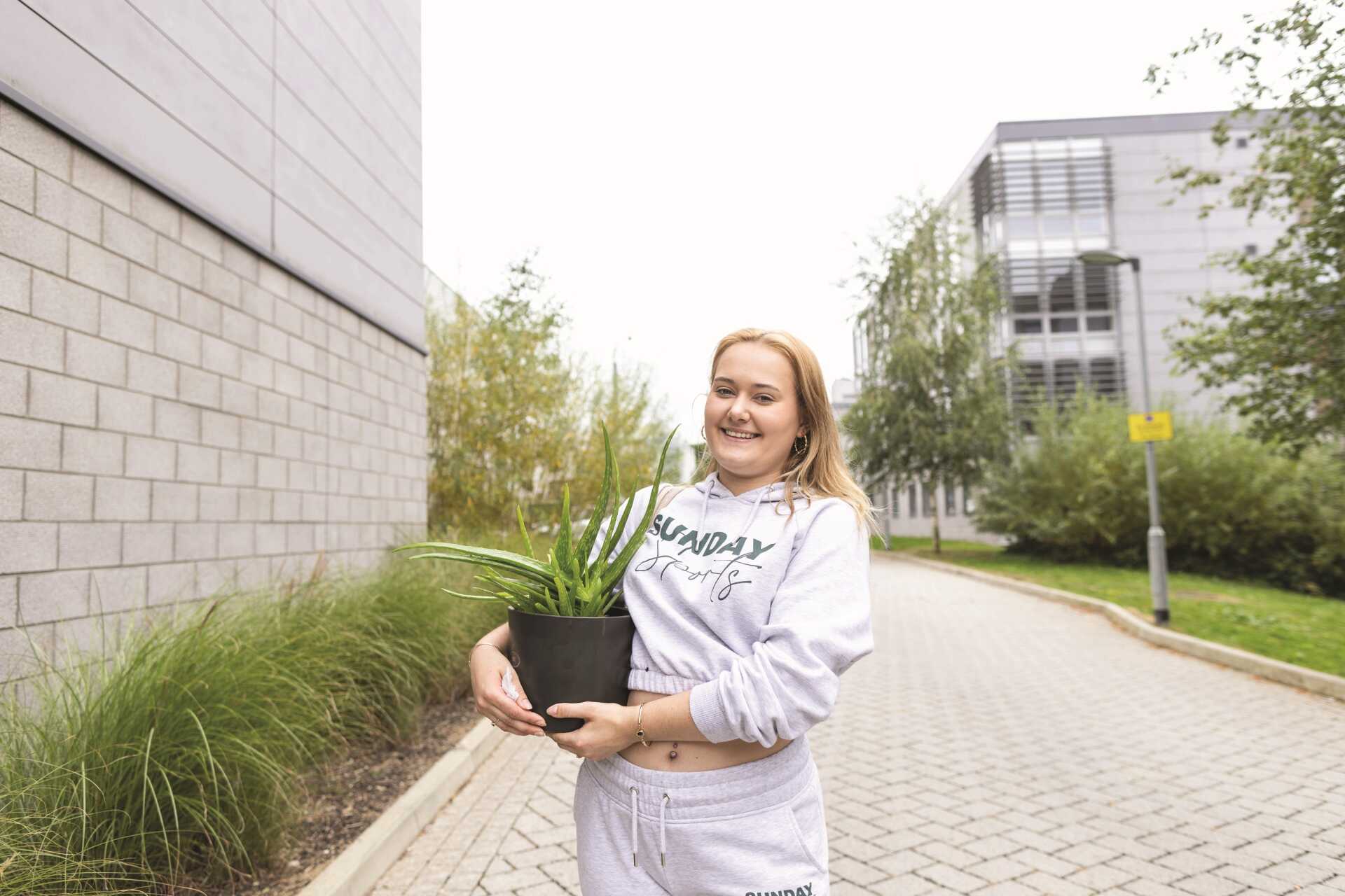 Female student smiling at camera,c carrying a plant when moving in to Turing College